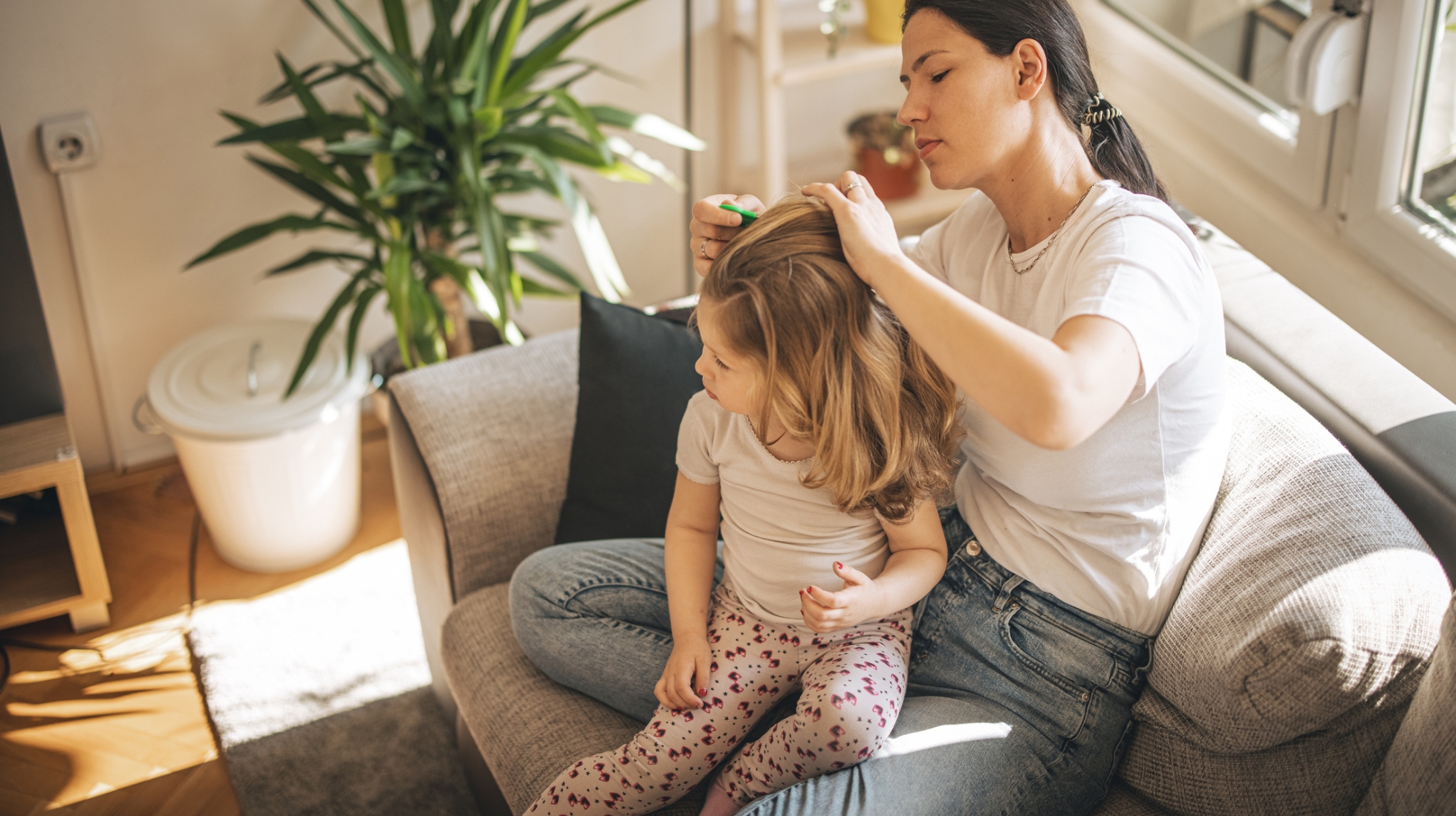 Äiti tutkii lapsen hiuksia ja etsii täitä sohvalla istuen. Taustalla viherkasi.