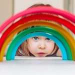beautiful-little-girl-looking-through-colorful-wooden-toy-rainbow-picture-id1196394004
