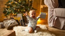 Mitä joululahjaksi yksivuotiaalle? Ota talteen käytännölliset ja hauskat lahjavinkit! Iloinen lapsi jouluna. Taustalla joulukuusi ja lahjoja.