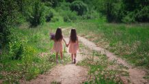 Backwards view of little identical twin sisters with long hair walking together holding hands at road among field in dresses at sunny nature in grass and flowers. Girls friendship and youth concept.