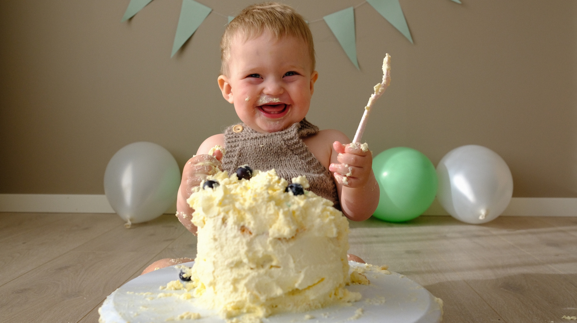 Iloinen taapero istuu lattialla ja syö lusikalla valtavaa kakkua. Taustalla juhlakoristeita.